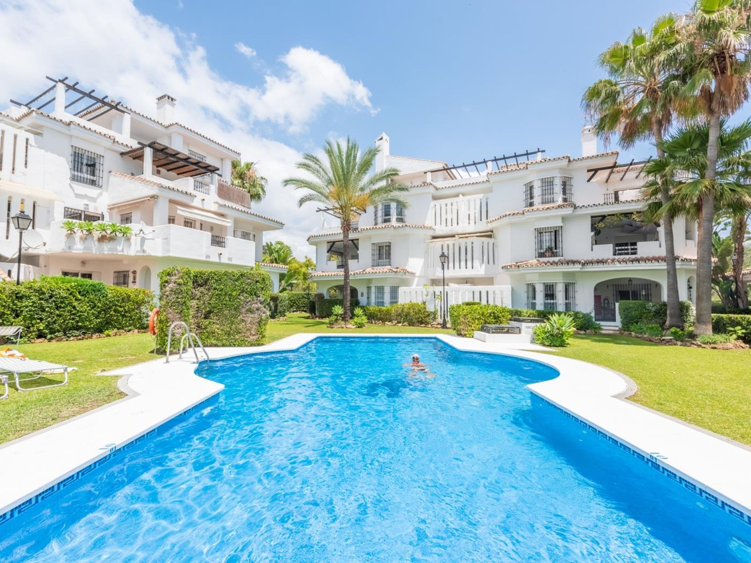 A swimming pool surrounded by multi-story buildings with balconies, a clear sky, and palm trees.
