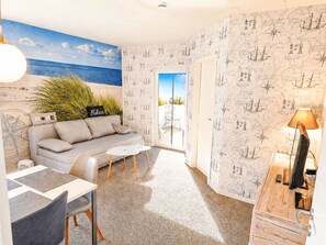 Ein modernes Wohnzimmer mit einer Couch, einem Couchtisch und Sesseln, Meerblick durch eine Glastür und nautisch thematischer Tapete.