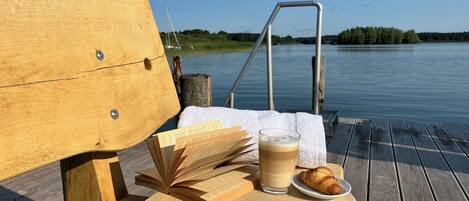 A wooden bench with a metal railing, a table with a book, a glass of beverage, and a croissant on a wooden deck by a calm body of water.