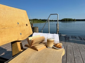 A wooden bench with a metal railing, a table with a book, a glass of beverage, and a croissant on a wooden deck by a calm body of water.