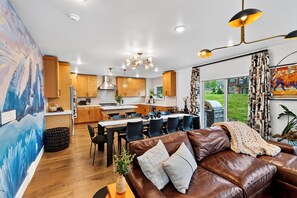 Eine moderne Küche mit Holzausstattung, ein Essbereich mit Tisch und Stühlen, ein Wohnzimmer mit Ledersofa und ein großes Fenster mit Blick ins Freie.