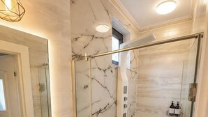 Ein Badezimmer mit einer gläsernen Duschkabine, einer Marmorwand, einem Waschbecken mit Pflanze und einem beleuchteten Spiegel.