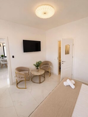 Ein modernes Hotelzimmer mit einem an der Wand befestigten Flachbildfernseher, einem Bett mit weißen Bettwäsche, zwei Sesseln, einem kleinen runden Tisch mit einer Pflanze und einer Tür zu einem Badezimmer.