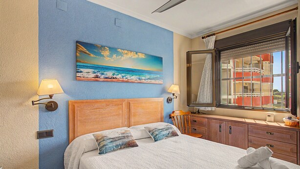 A bedroom with a bed, a painting of a beach scene, a window with a view of buildings, and a wooden headboard. - Casa Dolo · Beachfront Apartment with Pool, Elevator, and WiFi (Tavernes de la Valldigna)