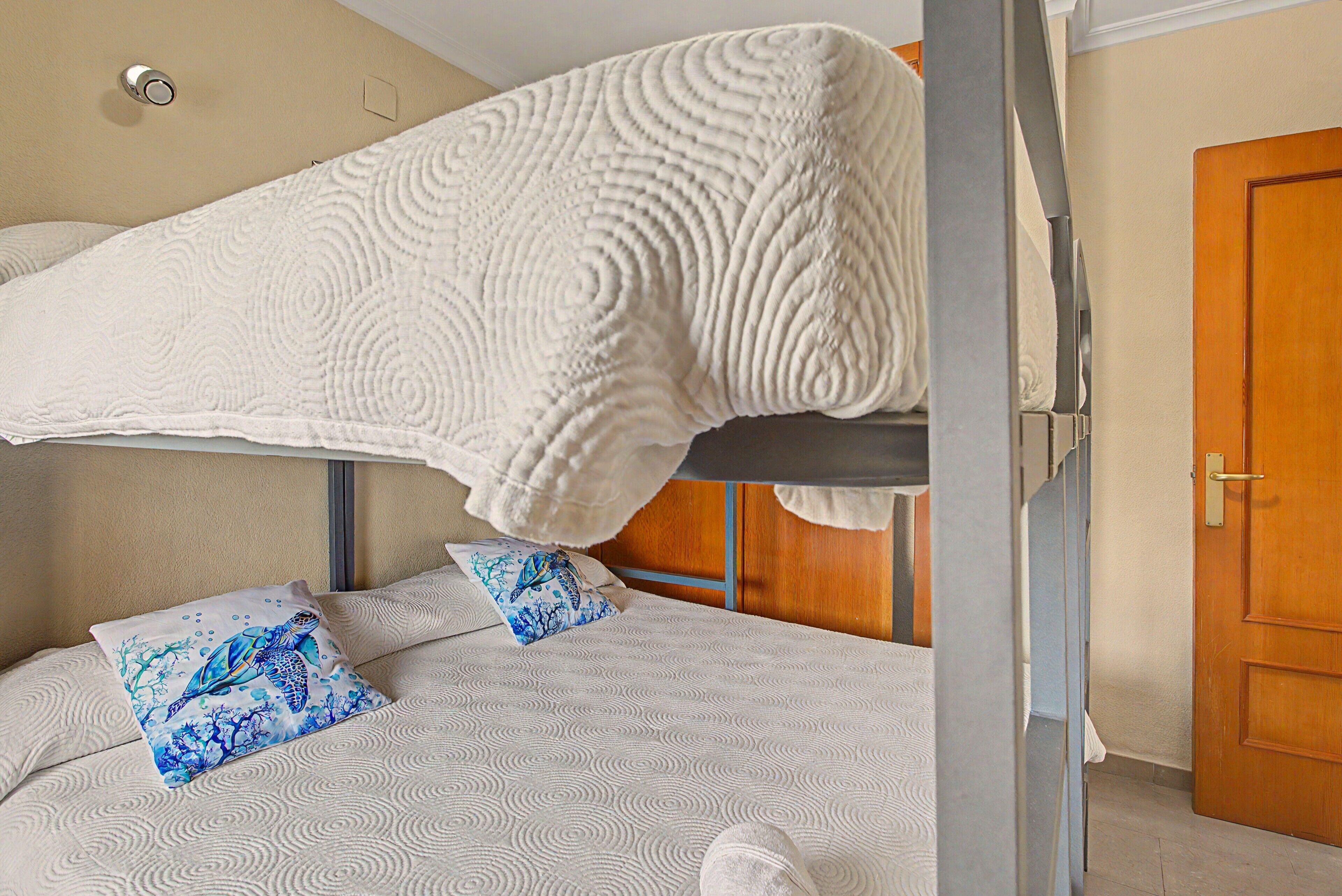 Un lit superposé recouvert d’une couverture blanche à texture, deux oreillers à motifs bleus et, au fond, une porte en bois.