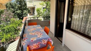 En balkon udendørs med et bord og stole, et vindue med gardiner samt udsigt til grøn vegetation.