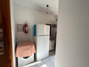 A compact kitchen with a washing machine, a refrigerator, and a fire extinguisher on the wall. - Özler Apart Pansiyon (Edremit)