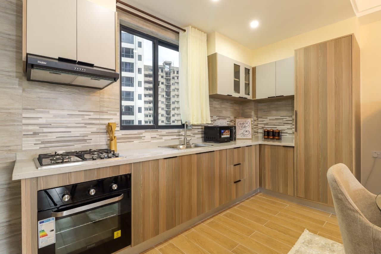 Una cocina moderna con estufa a gas, horno y microondas. Hay una ventana con cortinas, y la cocina cuenta con gabinetes de madera y un salpicadero de azulejos.