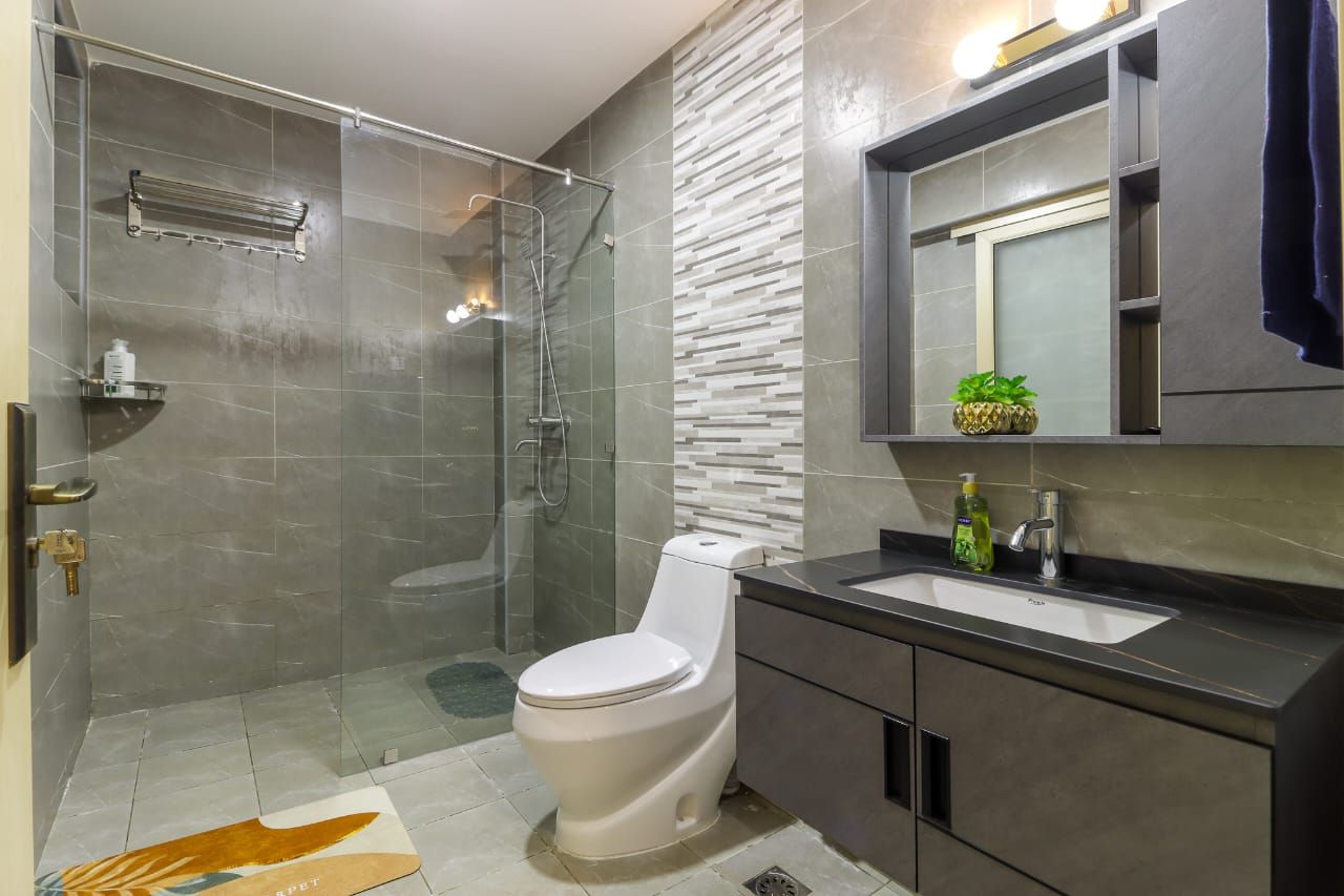 Un baño moderno con ducha envuelta en vidrio, inodoro blanco, tocador oscuro con lavamanos y espejo sobre el tocador.