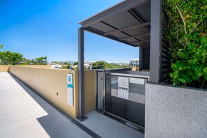 A covered outdoor area with a metal gate, a sign with emergency instructions, and a view of buildings in the distance. - Seashore Residences, Three-Bedroom 3B Deluxe (Thalang)