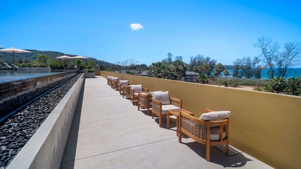 A balcony with a row of wooden chairs and a pool on one side. - Seashore Residences, Three-Bedroom 3A Premium (Thalang)