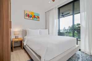 A hotel room with a large bed, a nightstand with a lamp, a painting on the wall, and a view of the outdoors through a window. - Seashore Residences, Three-Bedroom 3A Premium (Thalang)