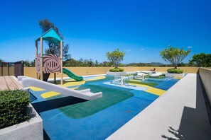 Ein Spielplatz mit Rutschen und Klettergerüsten, umgeben von einer Grünanlage mit Bäumen und blauem Himmel.