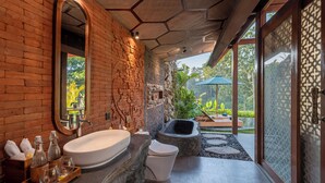 Baño con una bañera grande, un inodoro y un lavamanos. Hay una pared de piedra y una ventana con vistas a la vegetación.