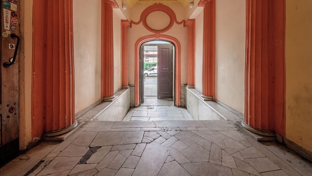 Un couloir au sol carrelé, orné de colonnes rouges, et une porte au fond.