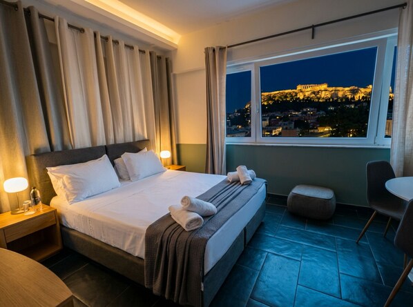 A hotel room with a bed, a nightstand, a chair, a table, and a window with a view of a cityscape at night. - Supreme Serenity Suite with Acropolis view - Located in the heart of Athens (Athina)