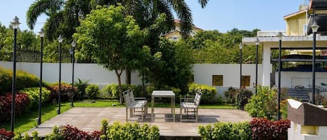A patio with a table and chairs, surrounded by lush greenery and a white wall.