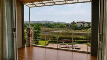 A room with a large glass door leading to a balcony with a view of a landscaped garden and distant hills.