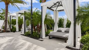 A modern outdoor lounge area with a canopy bed, palm trees, and a clear sky.