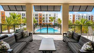 A modern lounge area with a pool view, featuring sofas, a coffee table, and a balcony with a view of a swimming pool and apartment buildings.