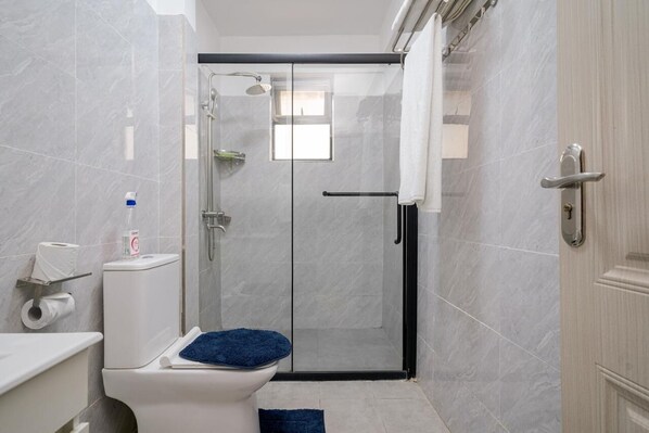 A modern bathroom with a glass-enclosed shower, a toilet with a blue mat, and a towel rack. - Diamond Homes View Palace Apartment (Nairobi)
