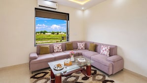 A living room with a sectional sofa, a glass coffee table, and a view of a green landscape through a large window. - Alaya Stays JSP Farm 4BHK Farmstay (Jaipur)