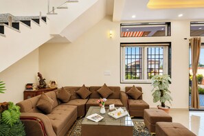 A modern living room with a sectional sofa, a coffee table, and a large window offering a view of the outside. - Alaya Stays JSP Farm 4BHK Farmstay (Jaipur)