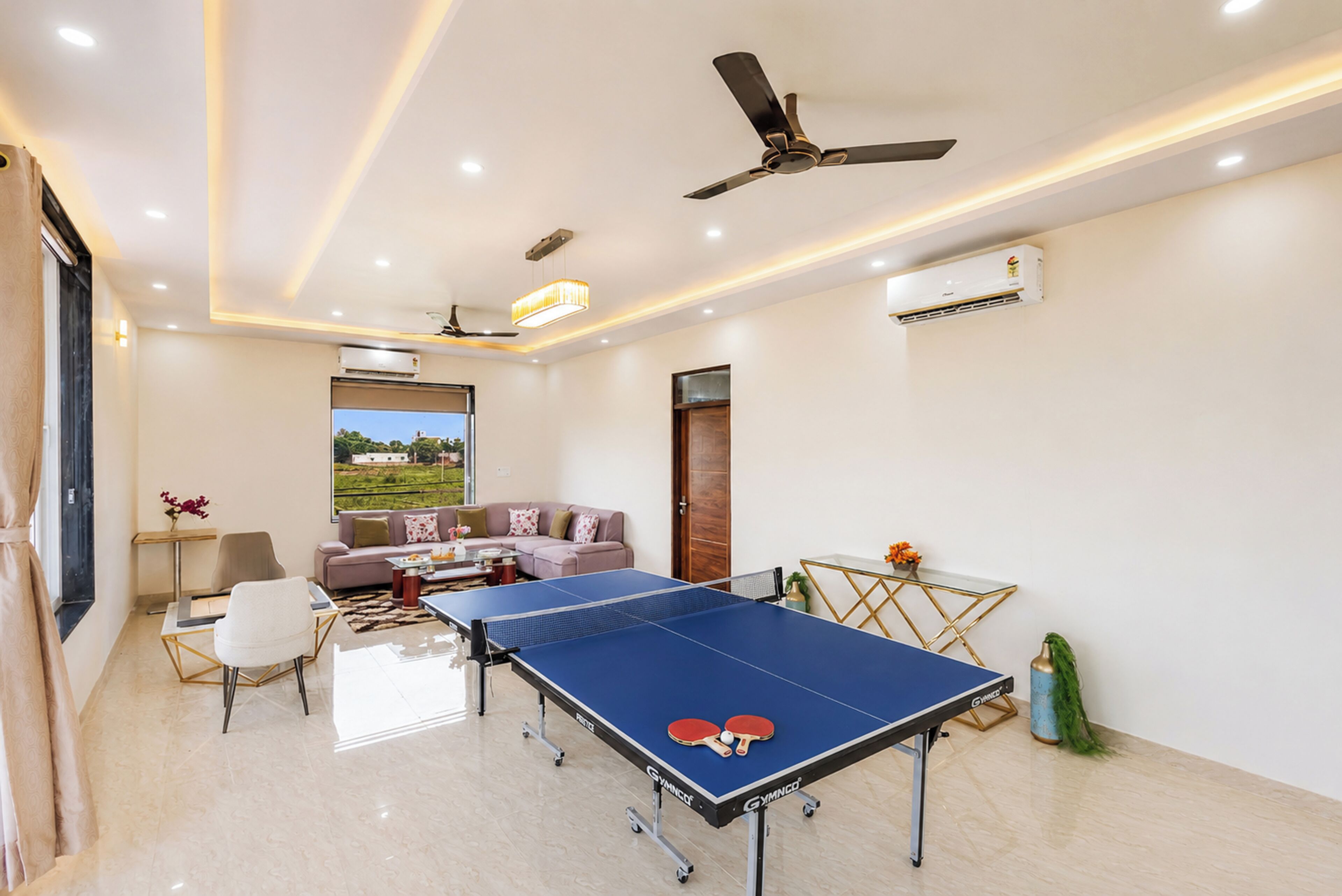 Una habitación con mesa de ping pong, ventilador de techo y vista a un paisaje verde a través de una ventana.