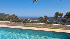 A swimming pool with clear blue water, surrounded by a stone wall and overlooking a coastal landscape with trees and a view of the sea. - Villa Vision | Brand New, Private Pool & Seaviews (Chania)