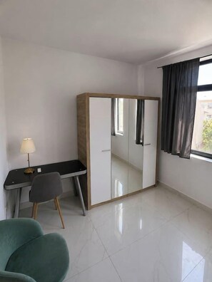 Ein Zimmer mit Schreibtisch, Stuhl, Kleiderschrank und einem Fenster mit Vorhängen.