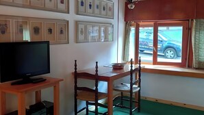 Een kamer met een televisie op een houten standaard, een eettafel met stoelen, een raam met uitzicht op een auto en ingelijste foto’s aan de muur.