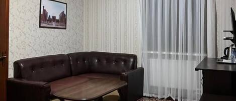 Ein Zimmer mit einer Couch, einem Tisch, einem gemusterten Teppich, einer Deckenleuchte und einem Vorhangfenster.