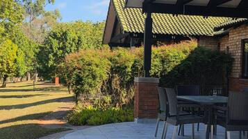 A covered outdoor seating area with a brick wall and a tiled floor, surrounded by greenery and trees.
