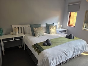 A neatly made bed with a green throw, a bedside table with a lamp, and a window with a view of greenery.