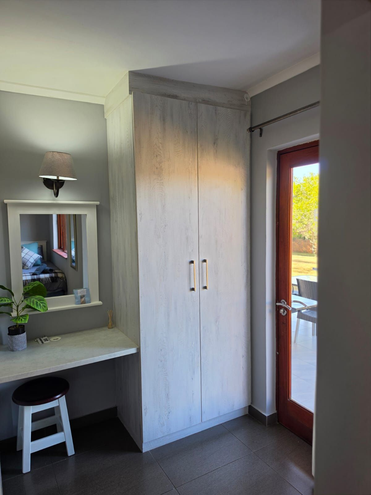 A room with a built-in desk, a mirror, a potted plant, a stool, and a wardrobe with two doors.
