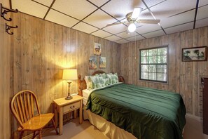 Una habitación con paneles de madera, una cama con cubrecama verde, una mesita de noche con lámpara, una silla y una ventana con persianas.