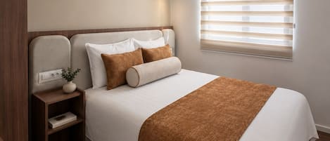 A modern bedroom with a large bed, a bedside table with a lamp, and a window with blinds.