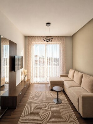 Ein modernes Wohnzimmer mit einer Couch, einem Fernseher, einem gemusterten Teppich und Blick durch transparente Vorhänge.
