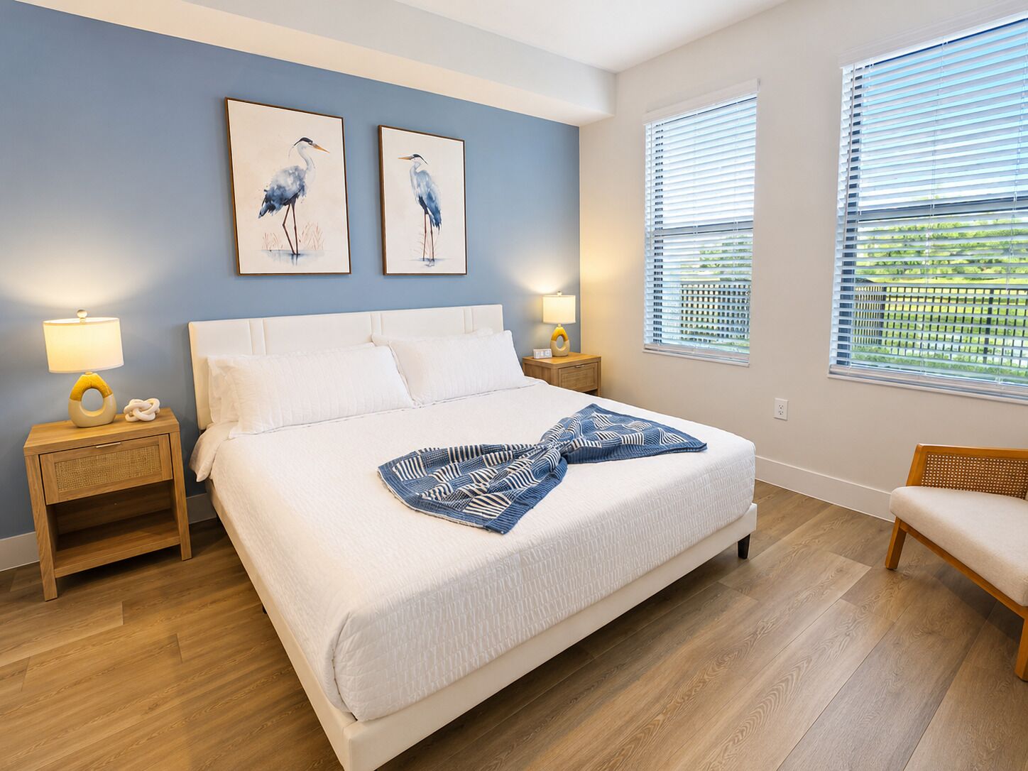A bedroom with a large bed, two bedside tables, a nightstand, a window with blinds, and two wall-mounted paintings of birds.