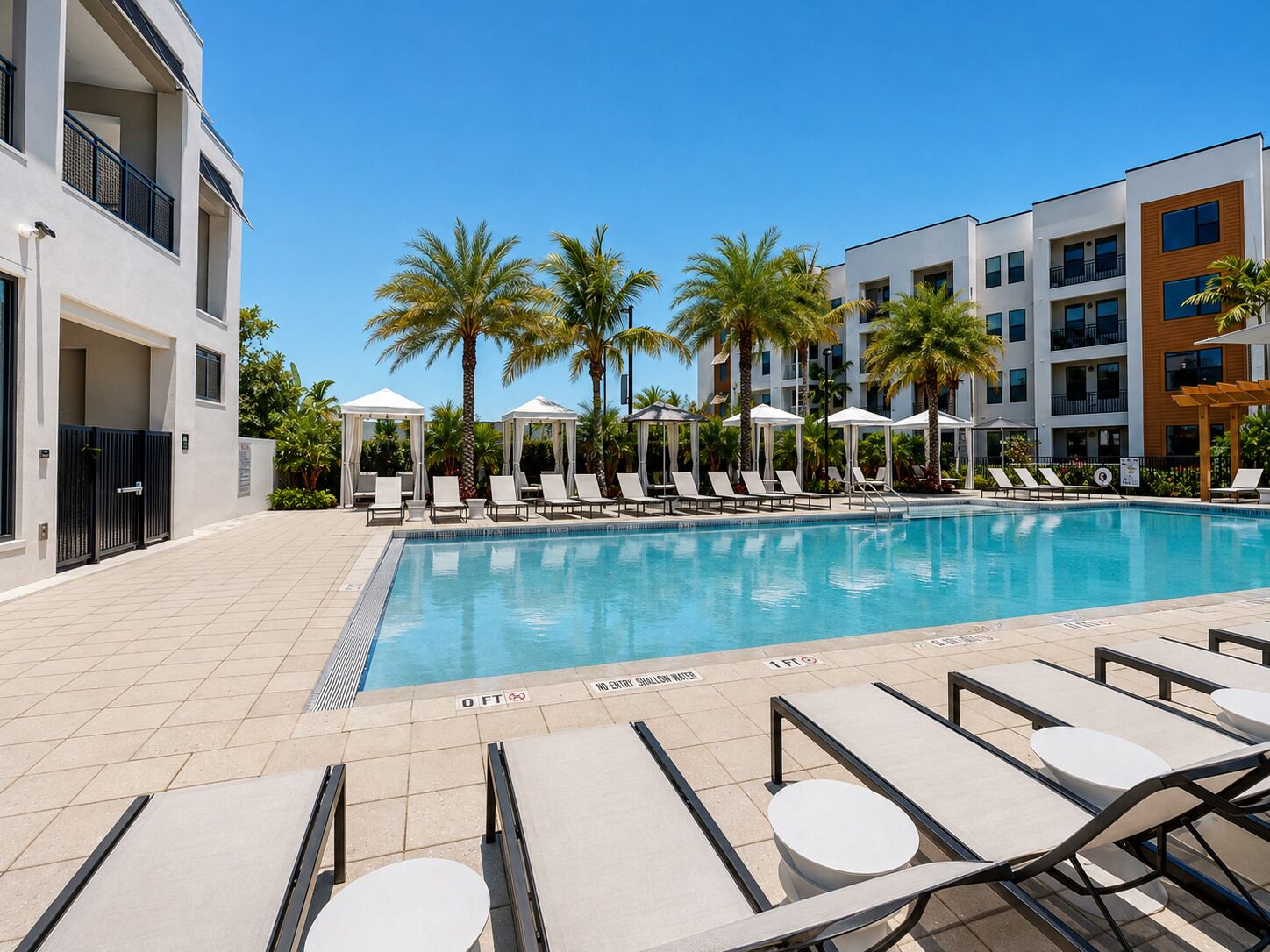 A swimming pool with lounge chairs and umbrellas, surrounded by palm trees and modern buildings.