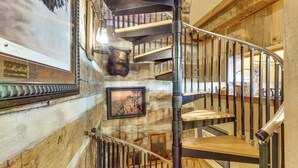 Una escalera de caracol con escalones de madera y barandillas de metal, que conduce a un rellano con un cuadro enmarco y una cabeza de animal montada.