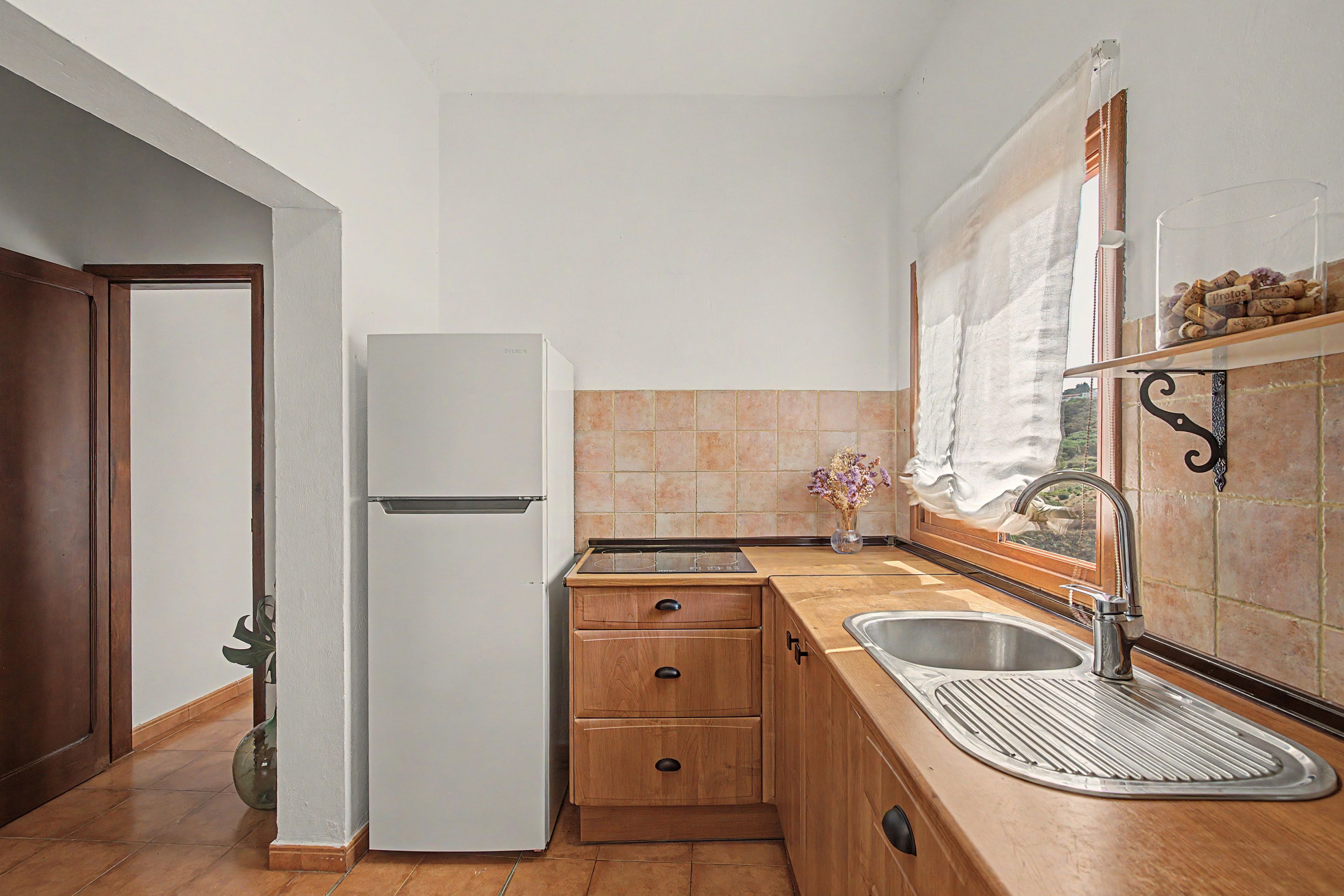 Una cocina compacta con refrigerador, gabinetes de madera, un lavamanos y un ventanal con cortina.