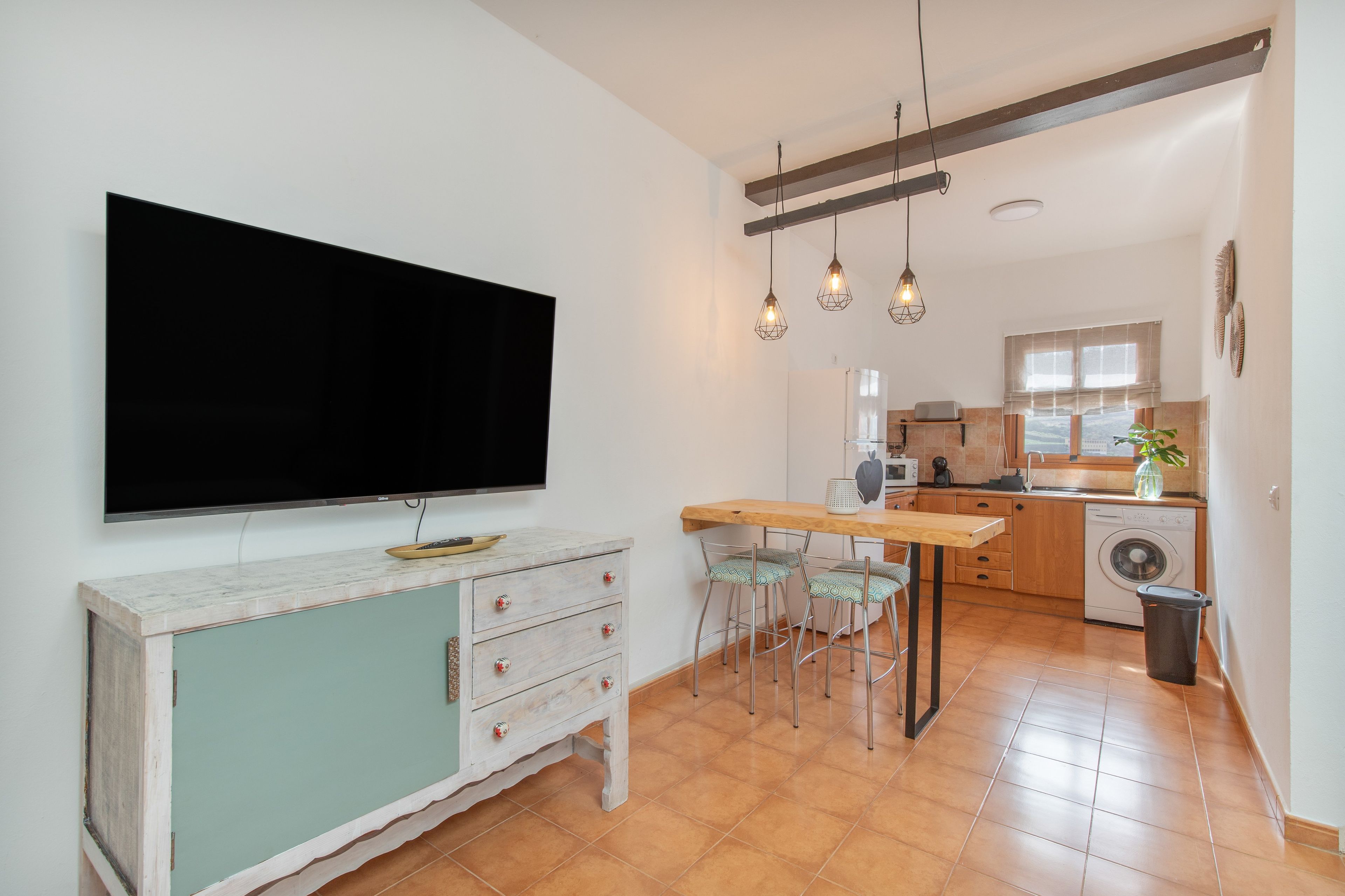 Una cocina moderna con televisor de pantalla plana, lámparas colgantes y un comedor con taburetes de bar.