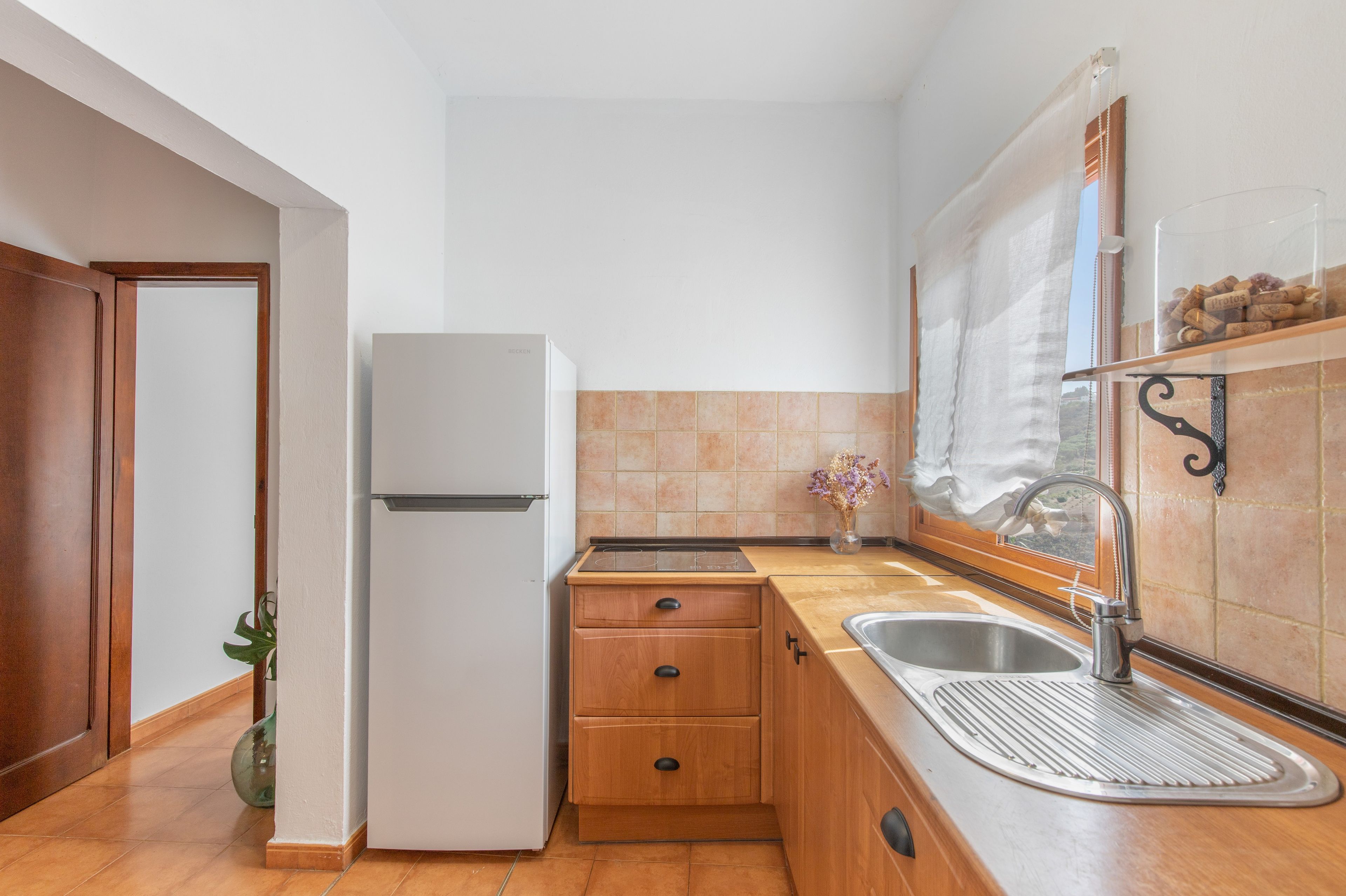 Een compacte keuken met een koelkast, houten kastjes, een spoelbak en een raam met gordijn.