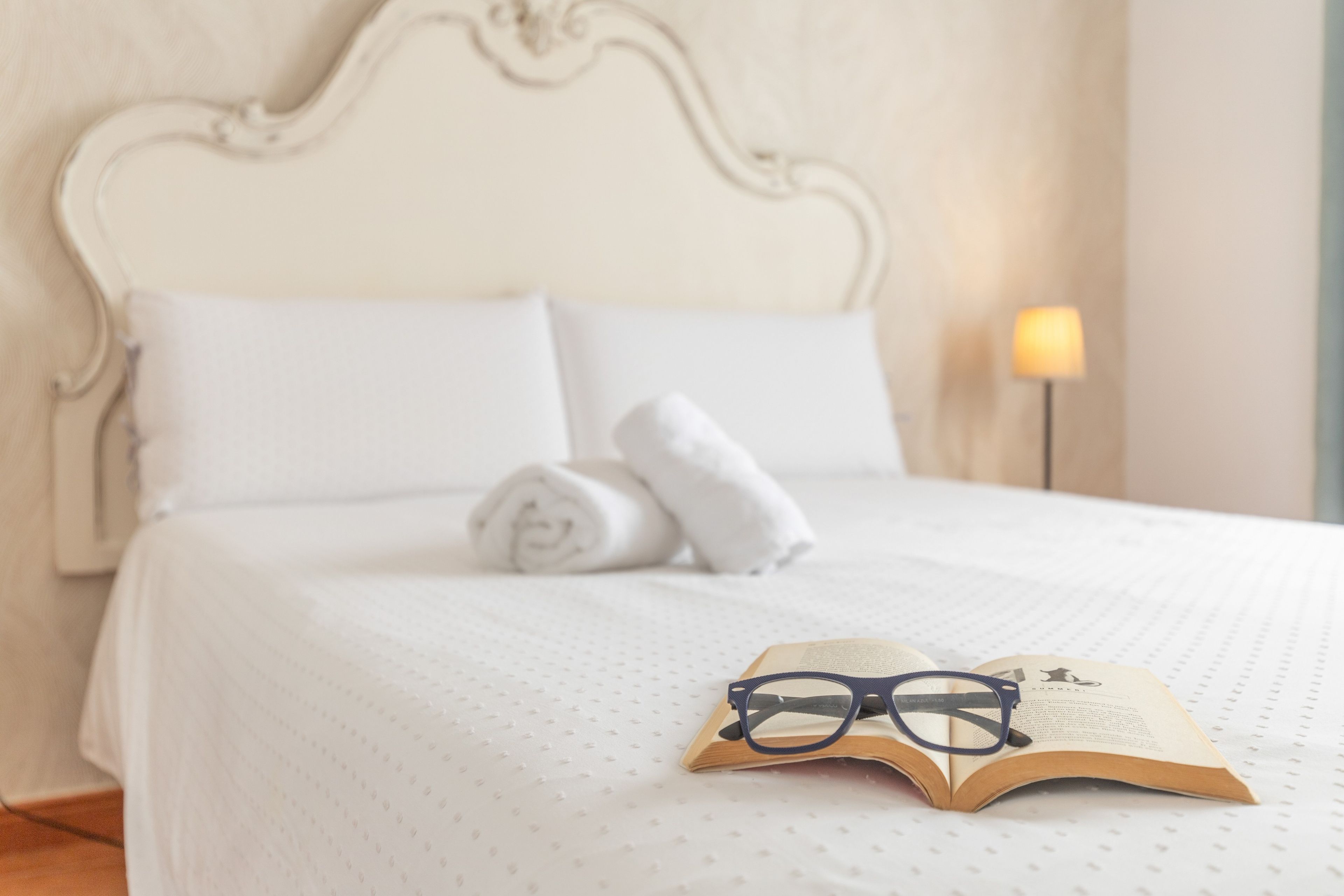 Een net opgemaakt bed met witte lakens, een opgerolde handdoek en een open boek met een bril erop.