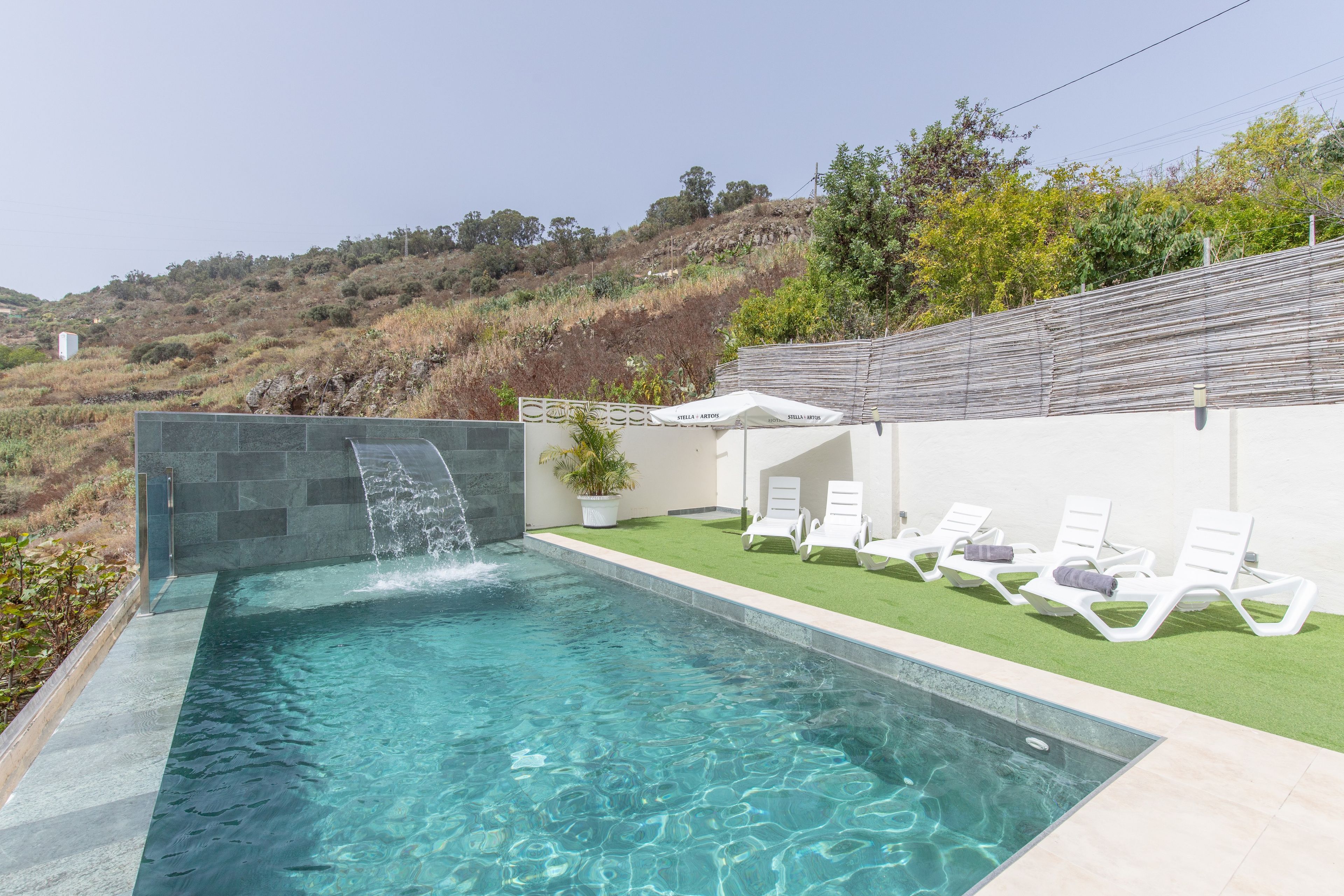 Una piscina con caída de agua, rodeada por un muro blanco y una zona de césped con sillas de descanso.