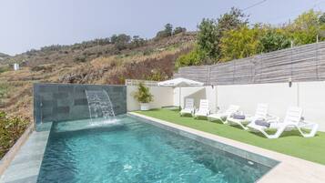 Una piscina con caída de agua, rodeada por un muro blanco y una zona de césped con sillas de descanso.