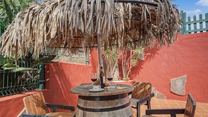 Ein Sitzbereich im Freien mit Holzstühlen und einem runden Tisch, ein Reetdach darüber und eine rote Wand im Hintergrund.