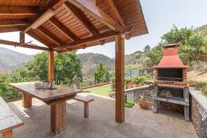 An outdoor patio with a wooden table and chairs, a barbecue grill, and a view of the surrounding landscape.