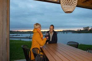 Two people seated at a wooden table outdoors with a view of a body of water and a bridge in the background. - Premium Water 4p Villa in Thorn (Thorn)